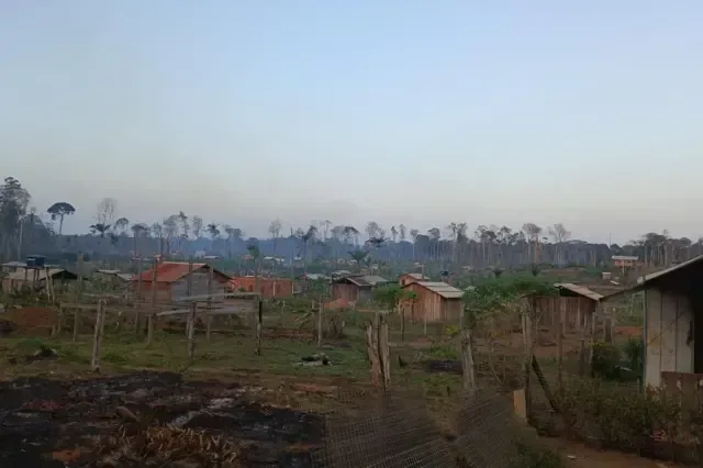 Área de ocupação rural na Amazônia, com casas simples de madeira espalhadas entre terrenos cultivados e cercas. Ao fundo, a vegetação foi amplamente desmatada, com árvores queimadas ou sem folhas e uma camada de fumaça sobre a paisagem.